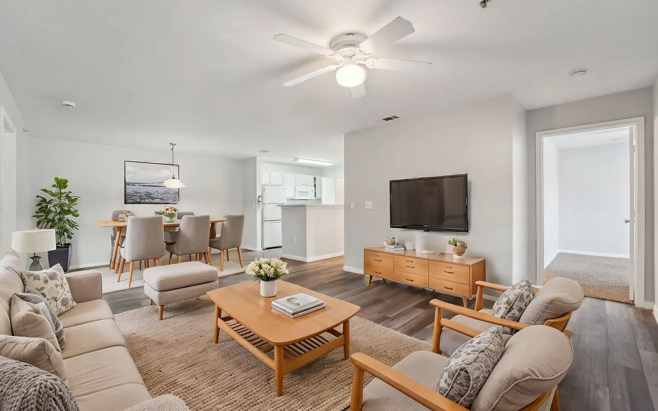 A modern living room featuring light-colored furniture, a wooden coffee table, and a flat-screen TV mounted on the wall. The decor includes a plant and a dining area visible in the background.