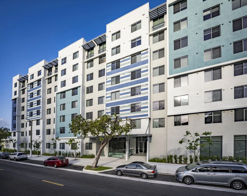 Contemporary Apartment Building A modern, six-story apartment building with a mix of white and light blue exterior panels, featuring large windows and green landscaping. Cars are parked along the street in front of the building.