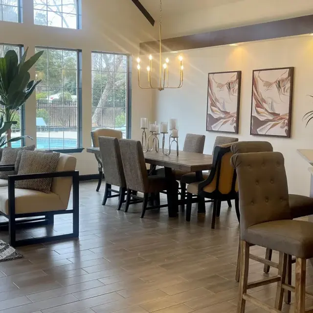 A bright and spacious living area with large windows, showcasing a dining table and chairs, modern seating, and decorative plants, with a view of a swimming pool outside.