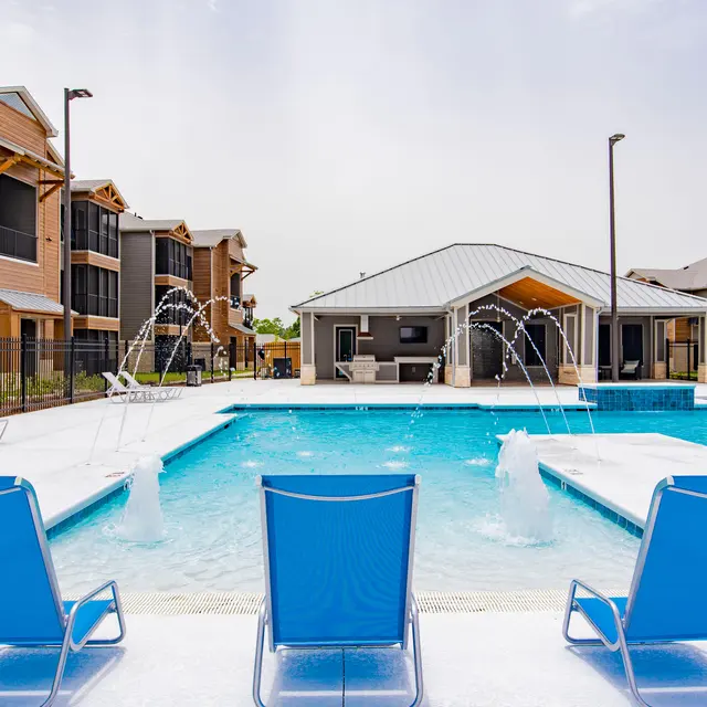 A sunny pool area with three blue lounge chairs in the foreground, surrounded by a sparkling blue pool with fountains. In the background, there are modern apartment buildings and a clubhouse with a grey roof.