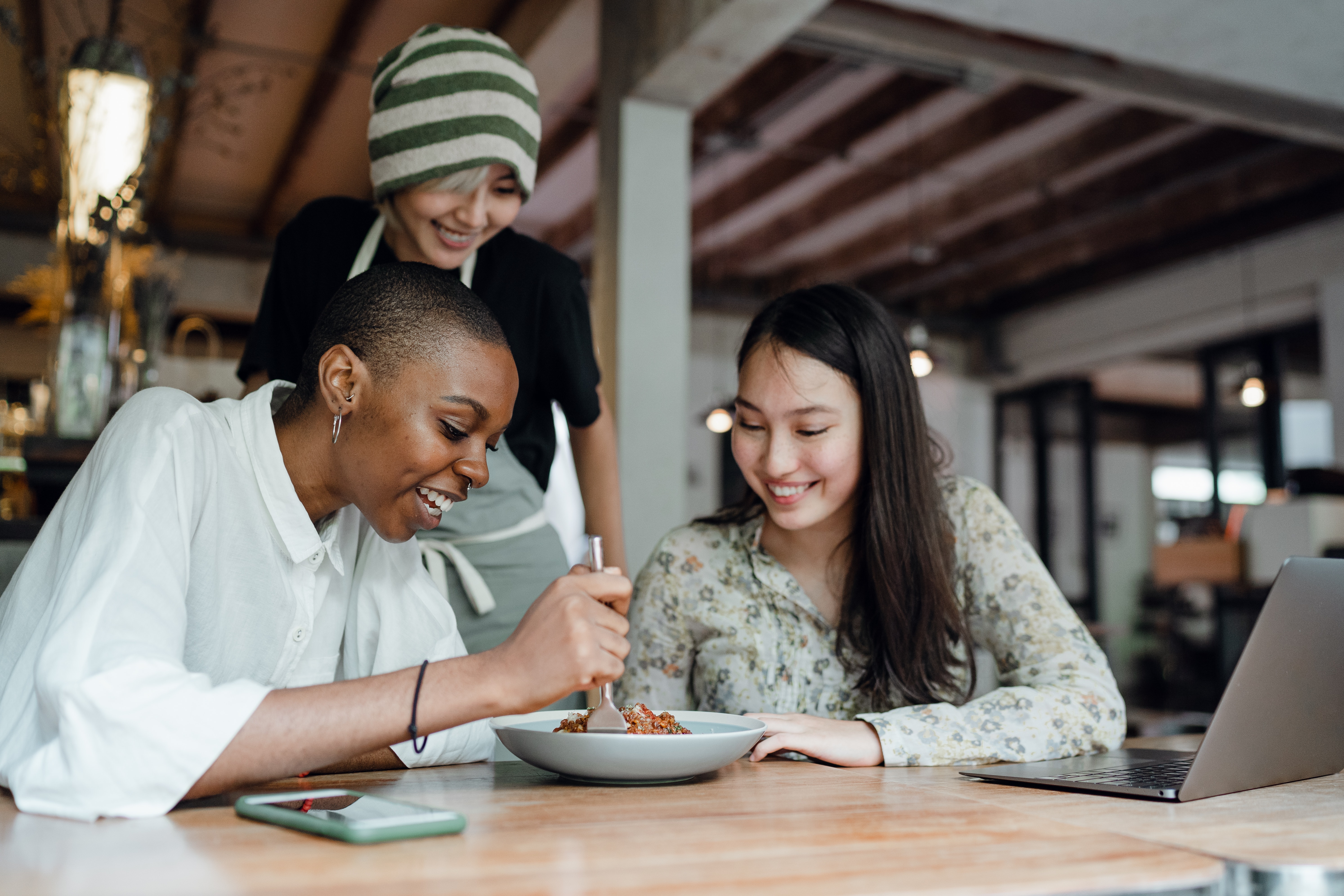Friends Sharing a Meal A group of three friends enjoying a meal together, with one person tasting food from a bowl while the others watch and smile. They are seated at a wooden table with a laptop and smartphone in front of them.