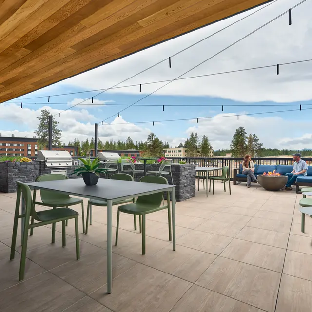 A spacious rooftop lounge area featuring wooden accents and a panoramic view. There are outdoor seating arrangements, including tables, chairs, and a fire pit, with greenery and string lights overhead. The sky is partly cloudy, and buildings and trees are visible in the background.