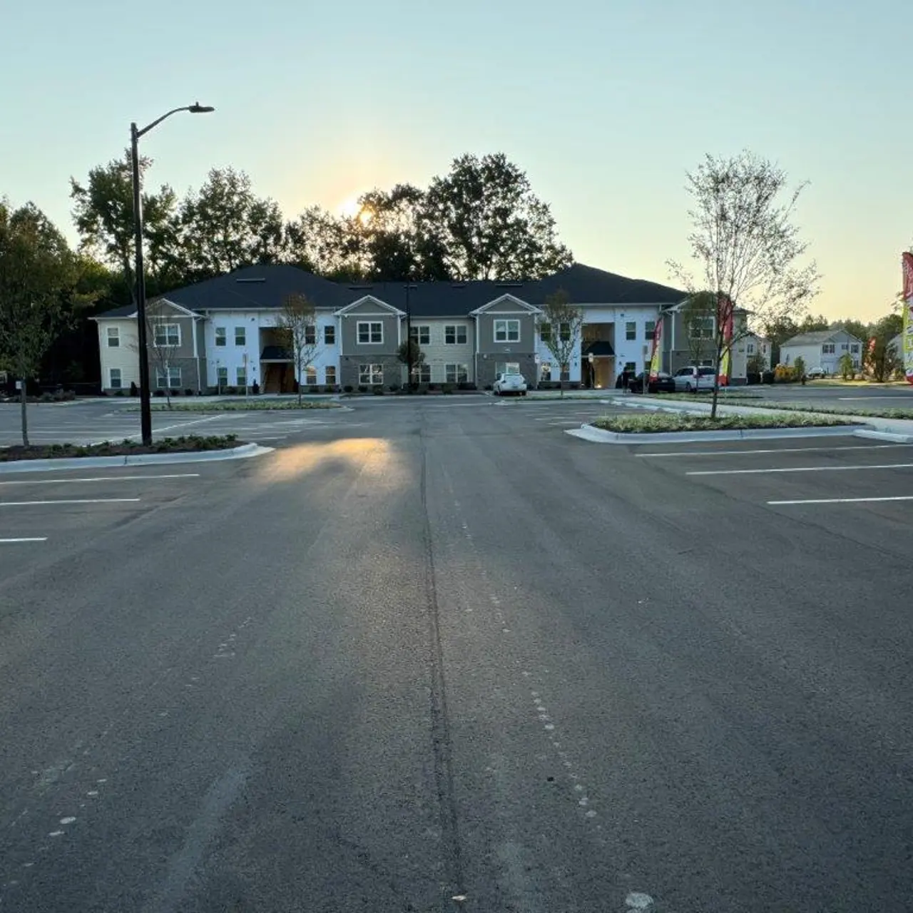 A view of an apartment complex from the entrance of a large parking lot. The sun is rising in the background, casting a warm light. There are several parked cars and a landscaped area in front of the buildings.