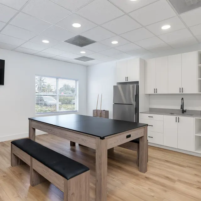 A well-lit common area featuring a large wooden table with a black surface, a bench attached on one side, and a modern kitchen setup in the background with a refrigerator and cabinets. There is a blue bean bag chair on the left and large windows letting in natural light.