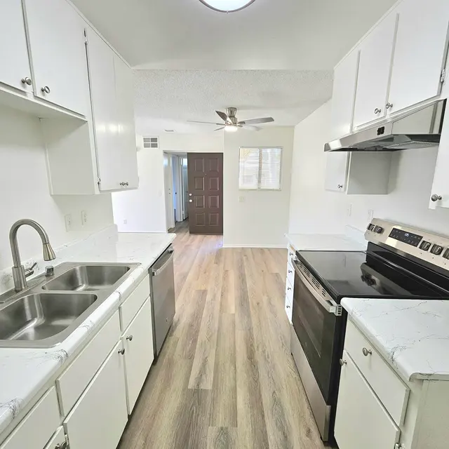 A modern kitchen featuring white cabinetry, stainless steel appliances, and wooden flooring. Two sinks are visible on the left side, with a view of an open layout leading to a door.