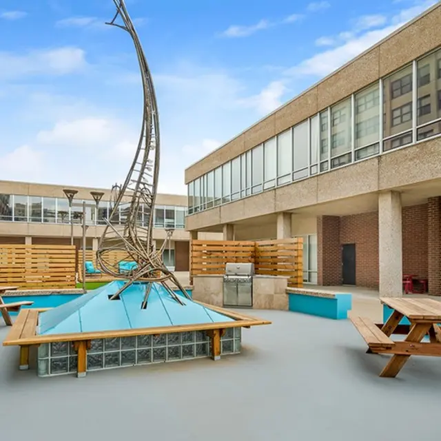 A modern courtyard featuring an artistic metal sculpture, picnic tables, and blue pool areas surrounded by buildings.