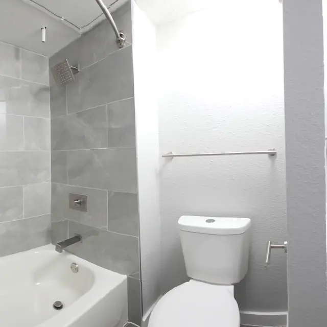 A modern bathroom featuring a bathtub and shower on the left, and a sink and toilet on the right. The walls are tiled with light gray porcelain tiles, and there is a round mirror above the sink.