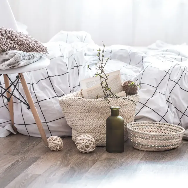 Cozy bedroom scene with a white bedspread, woven baskets, a plant, and decorative items on wooden flooring. Soft lighting.