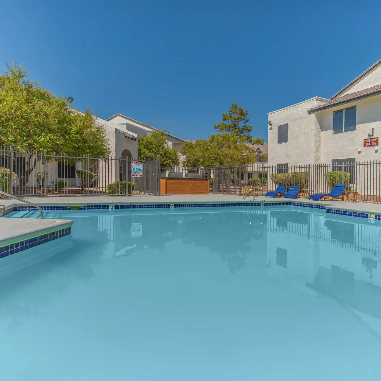 A clear blue swimming pool surrounded by a fenced area with green shrubs and trees. Two blue lounge chairs are visible next to the pool, with white buildings in the background.