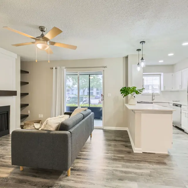 A modern living area featuring a gray couch, a fireplace, and an open kitchen layout with white cabinets and stainless steel appliances, creating a bright and inviting atmosphere.