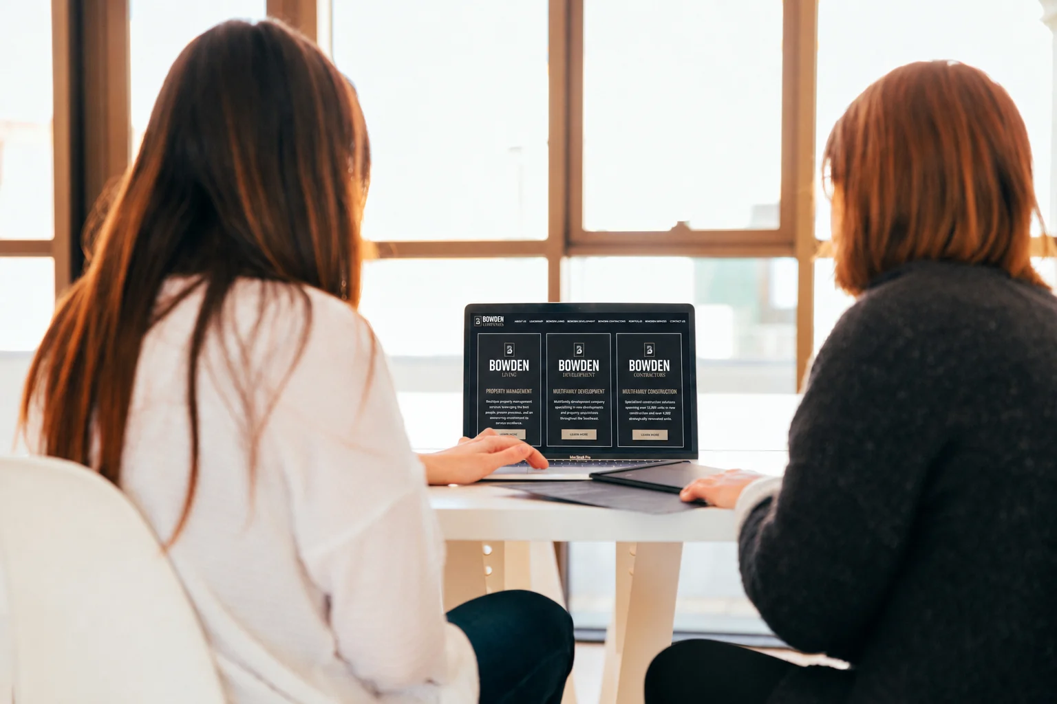Women Working Together in an Office Two women seated at a table looking at a laptop screen in a modern office environment with large windows.