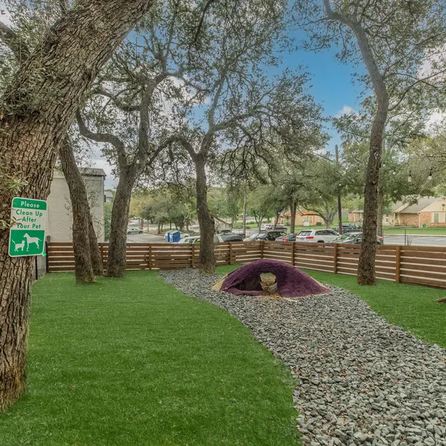 Artisan at South Lamar Dog park with grass and gravel walkway featuring a tunnel for pets to walk through and enjoy along with multiple tree's.