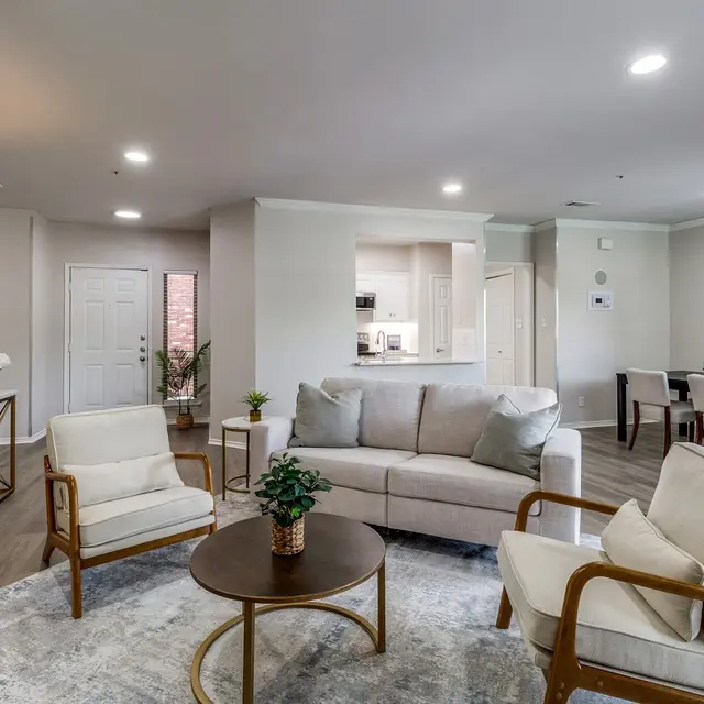 Modern Living Room Interior A modern living room featuring a light grey sofa, two white armchairs, and a round coffee table on a patterned rug. There is a dining area visible in the background, with a table and chairs. The room has soft lighting and neutral walls, decorated with a minimalistic art piece.