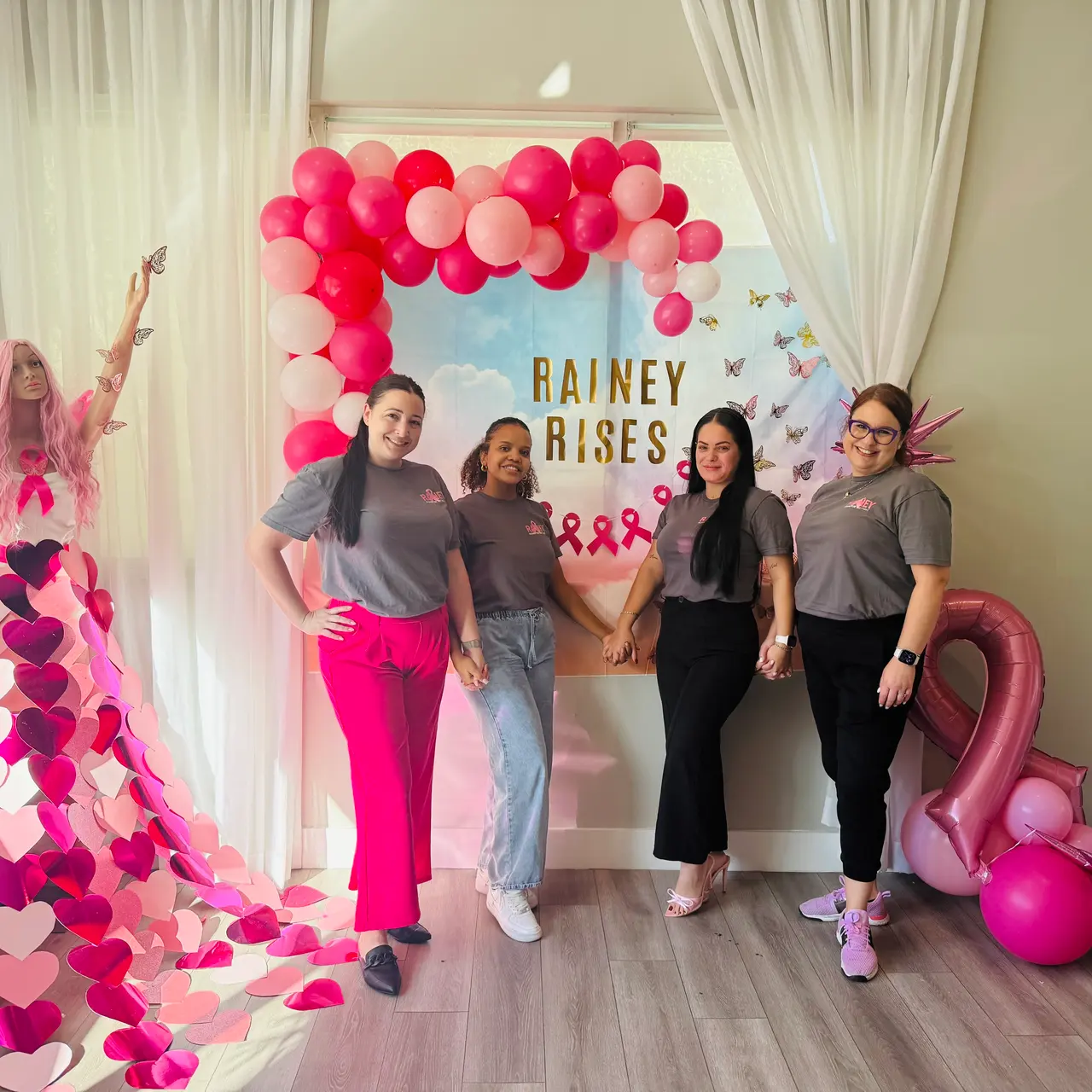 A group of five women standing together for a photo at an event. They are in front of a backdrop with the text 'Rainey Rises' surrounded by pink and colorful balloons. One woman on the left is wearing a costume made of pink hearts. The setting is bright and festive.