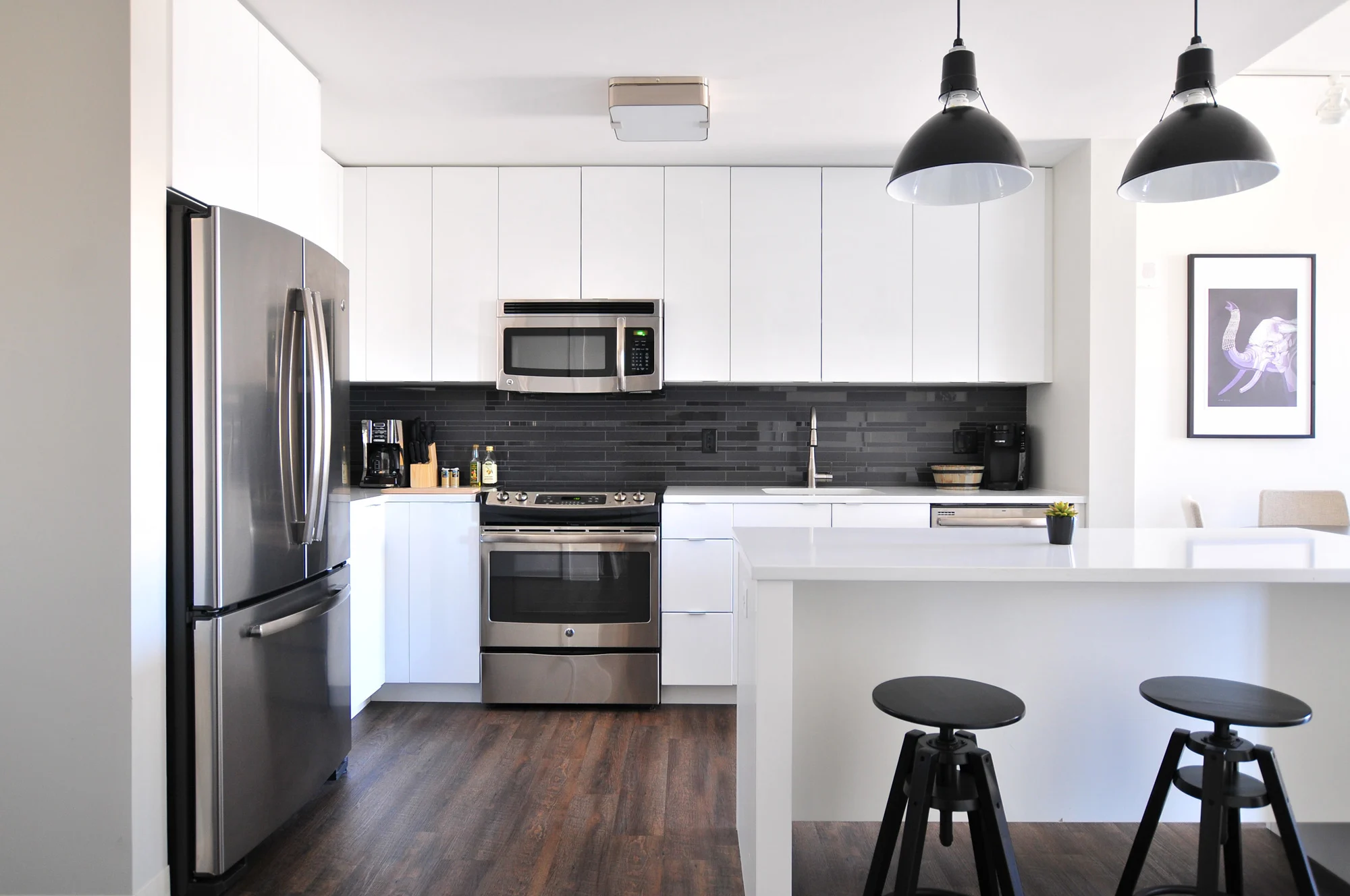 Premium Apartments Modern kitchen featuring stainless steel appliances, white cabinets, dark backsplash, and black stools at a sleek island.