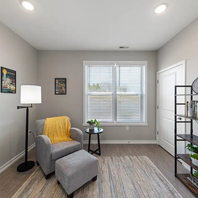 The Links at Franklin Franklin IN Apartments - The Links at Franklin - Corner Side Room with Wood-Style Plank Flooring, Area Rug, Grey Armchair, Ottoman, Carpet, Storage Rack with Decorations and Plants, Large Window, and Framed Art
