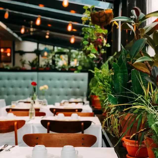 Charming Restaurant Interior with Greenery A cozy restaurant interior featuring a green tufted seating area, white tablecloths on wooden tables, and vibrant indoor plants.