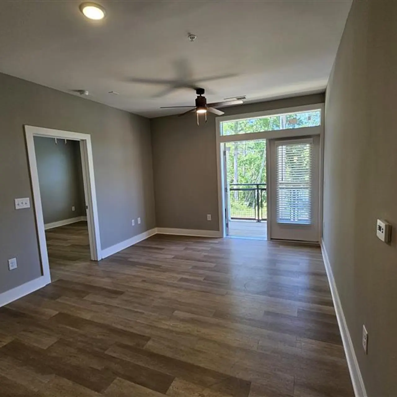 A spacious living room with light brown wooden flooring, gray walls, a ceiling fan, and a balcony door leading outside. There is a doorway to an adjacent room on the left.