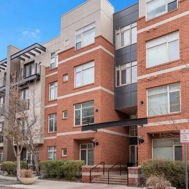 A modern apartment building featuring a mix of brick and stucco facades, with balconies and large windows. The entrance is well-defined with stairs leading up to the doors.