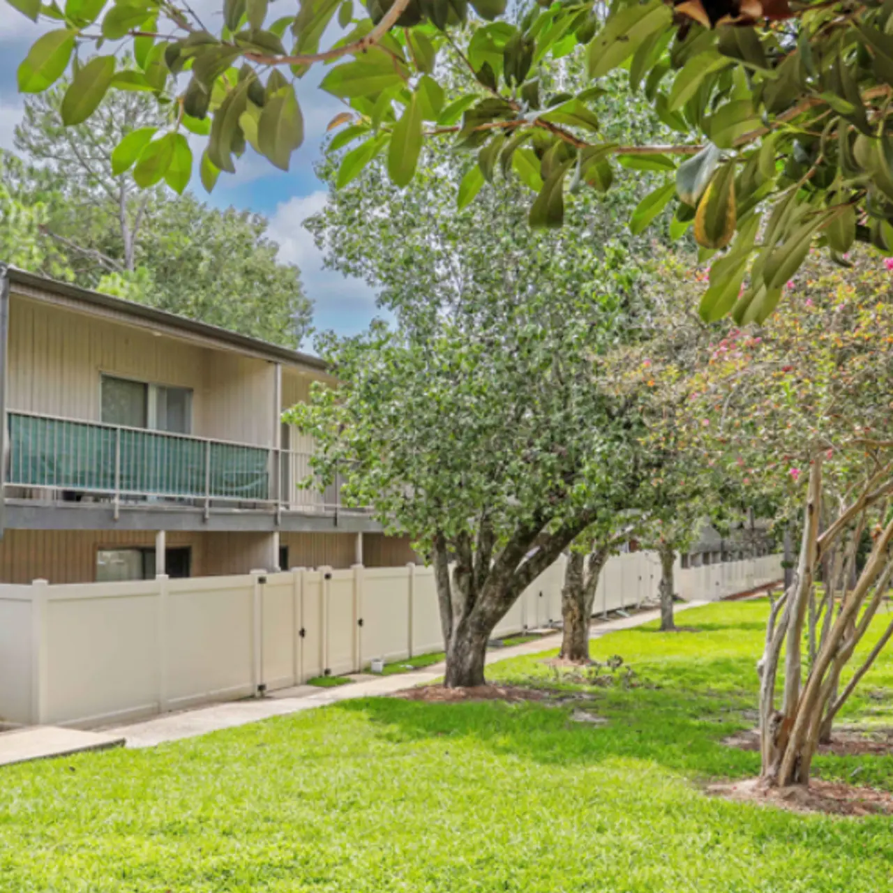 A view of an apartment complex with two stories, surrounded by green grass and trees, under a partly cloudy sky.