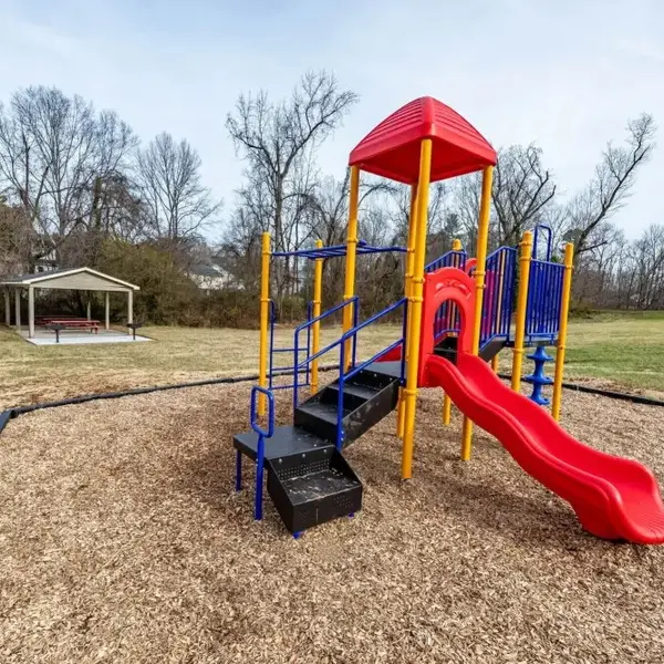 A colorful playground featuring a red slide and climbing structure, surrounded by wood chips, with a gazebo in the background and trees in the distance.