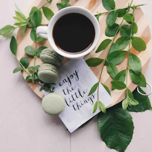 College Crest A cup of coffee on a wooden plate surrounded by green leaves and macarons, with a note that says "enjoy the little things."
