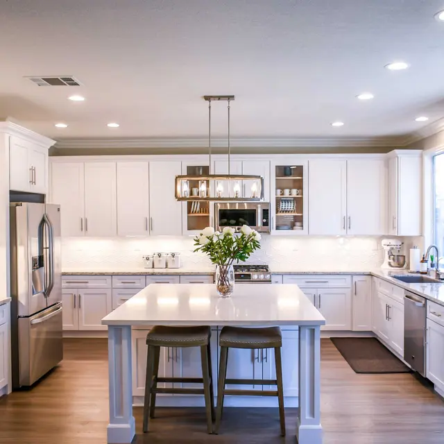 Modern kitchen featuring white cabinets, a large island with two bar stools, stainless steel appliances, and bright lighting.