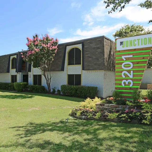 Exterior view of a multi-unit apartment building with a vibrant sign that reads 'The Junction 3201'. The building features a modern design with landscaped greenery and trees in the foreground.