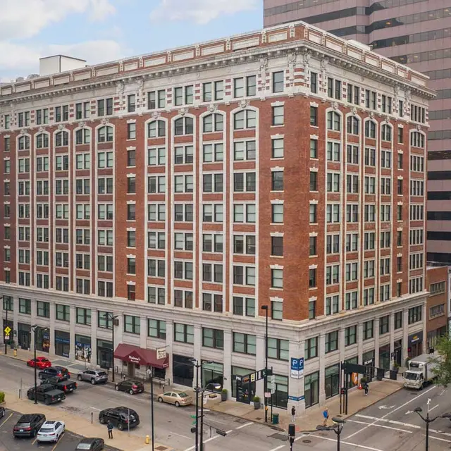 A multi-story red-brick building with white accents, surrounded by city streets and parked cars, adjacent to a floral mural on the left.
