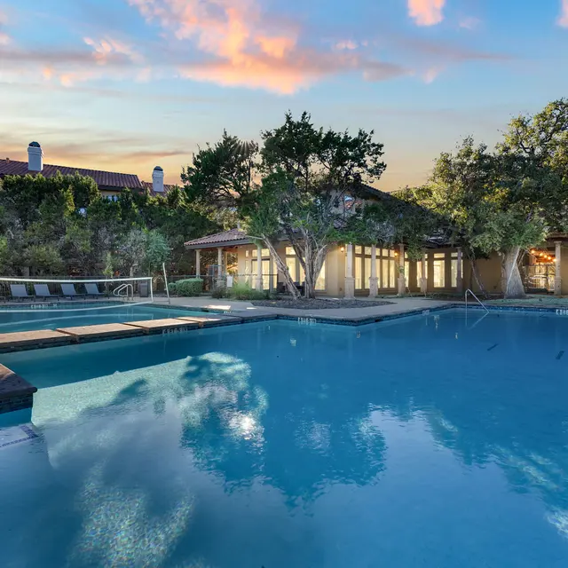 A calm swimming pool surrounded by trees and a building in the background during sunset, with vibrant orange and blue hues in the sky.