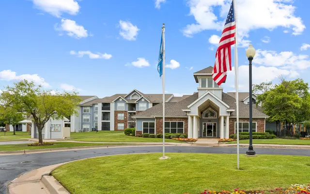 A residential building with large windows, surrounded by greenery, flags, and colorful flowers under a partly cloudy sky.