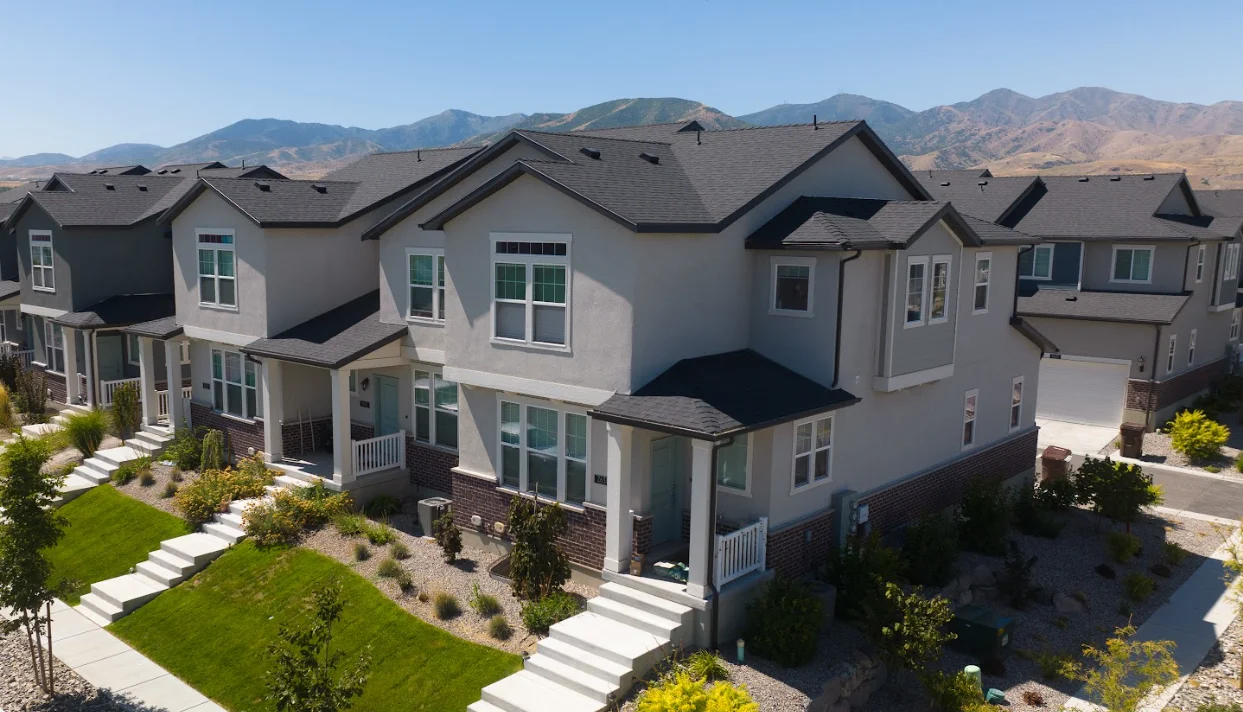 Aerial view of a modern housing development with several two-story homes, green lawns, and pathways, set against a mountainous background.