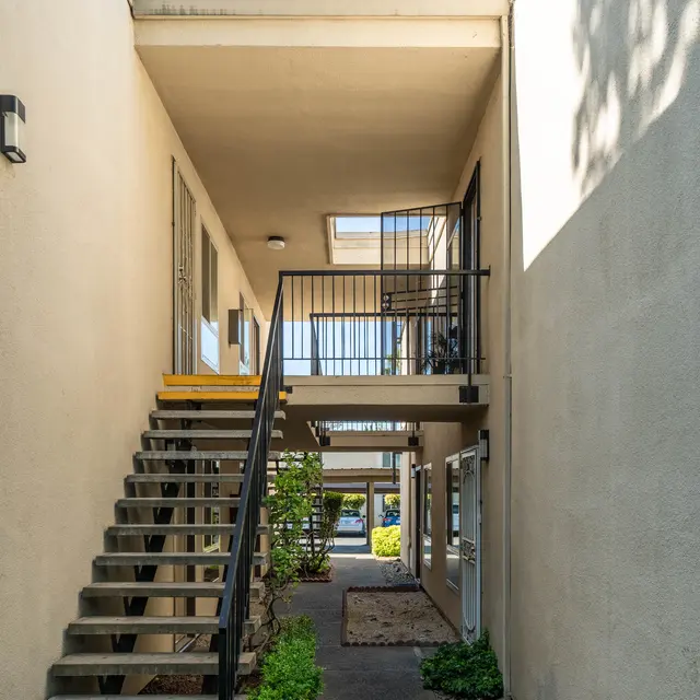 A view of an apartment complex from the entryway, featuring stairs leading to a second level and greenery around the pathway.