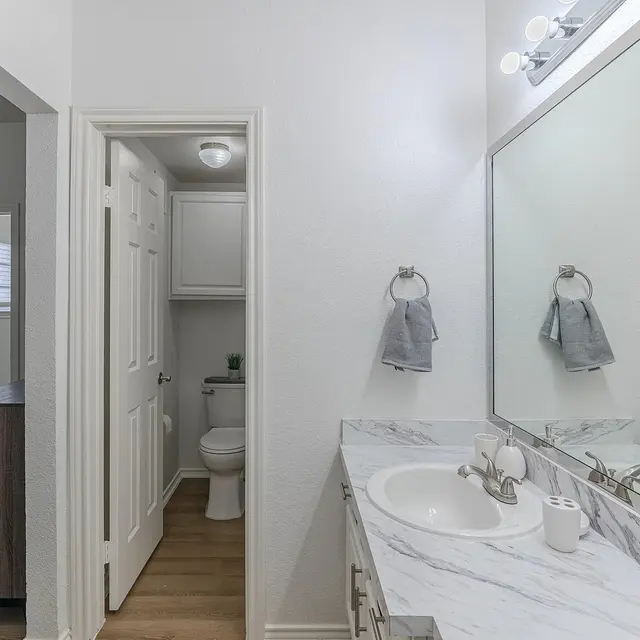 A spacious modern bathroom featuring a double vanity with a marble countertop, grey towels hanging by the sink, and a light fixture above the mirror. There is a door leading to a toilet area visible in the background.