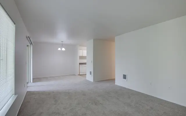 A spacious living room with light-colored carpet and white walls, featuring a large window with blinds and a doorway leading to a kitchen area. There are two light fixtures hanging from the ceiling.