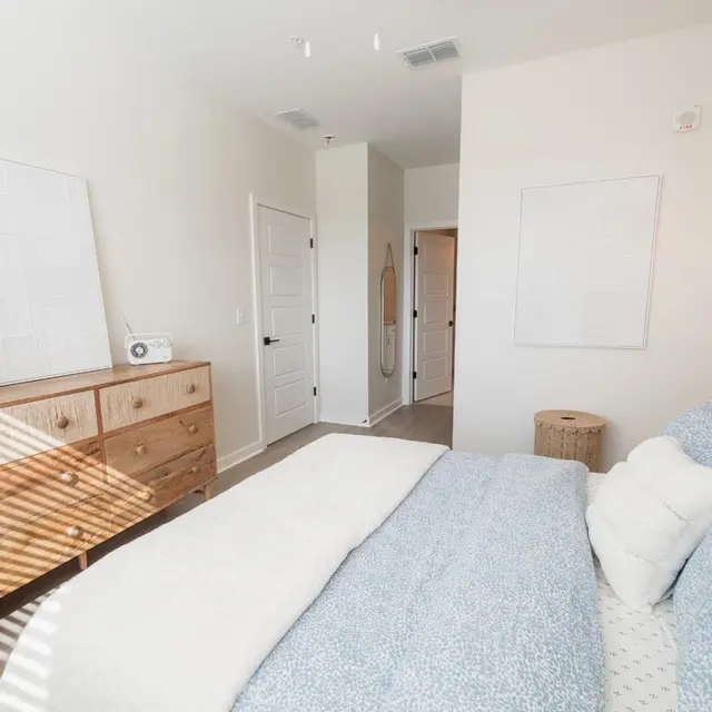A modern bedroom featuring a queen-size bed with light blue bedding, a wooden dresser with a vase of dried flowers, and a bedside lamp. Sunlight casts shadows across the room, highlighting the minimalist decor.