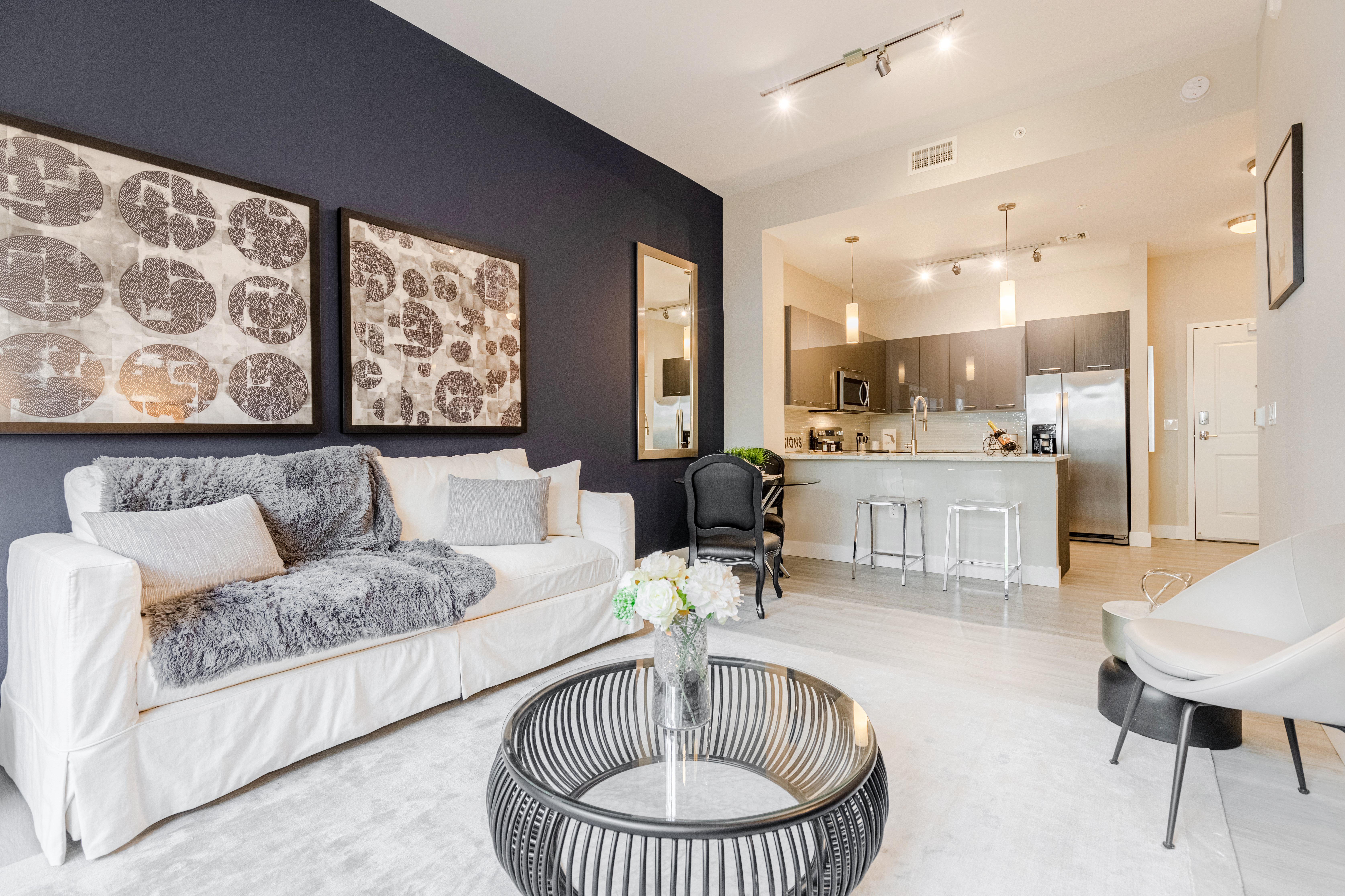Modern Living Room Design A modern living room featuring a white couch with gray throw, abstract artworks on the wall, and a black circular coffee table. The room has a dark blue accent wall and a kitchen area in the background.