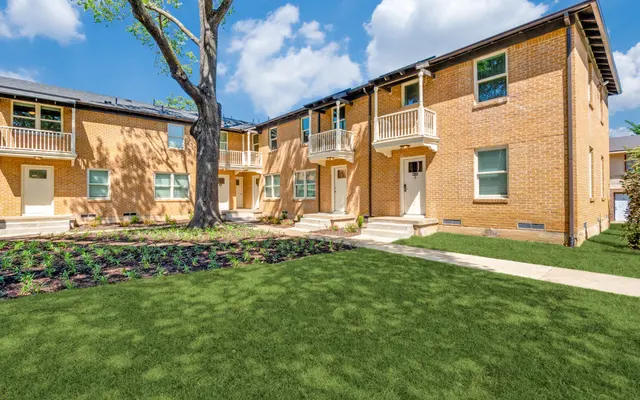 Exterior view of a two-story brick apartment complex with a green lawn and a large tree in the front.