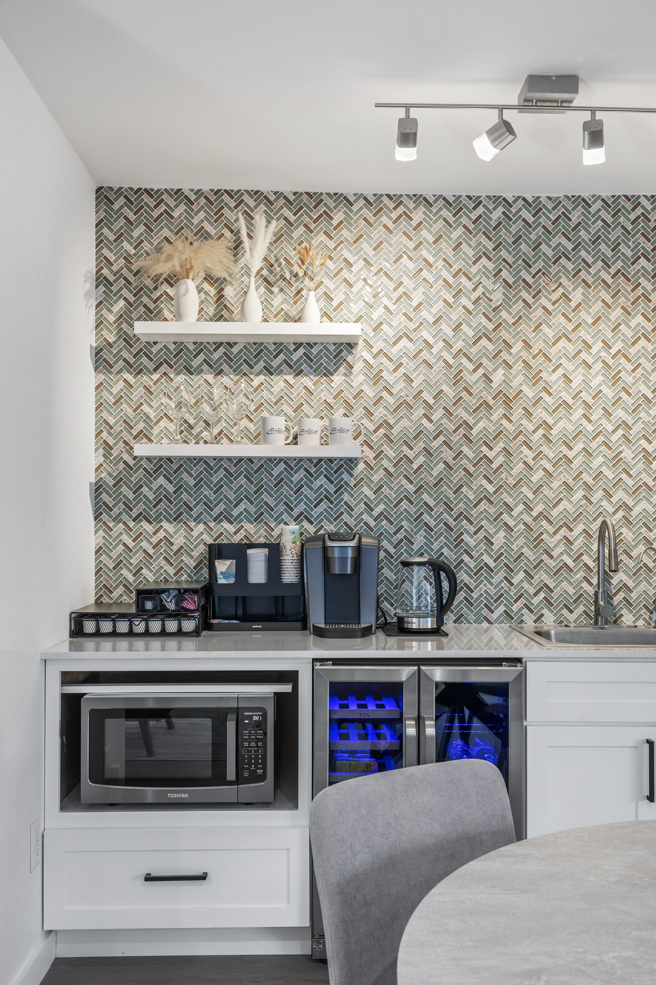A modern kitchen featuring a patterned feature wall, open shelves with decorative items, a coffee maker, kettle, microwave, and a small fridge with a blue interior light. There's a round table with a grey chair in the foreground.
