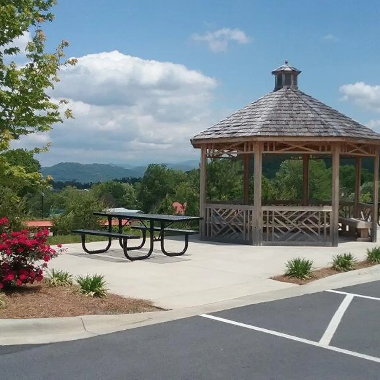 A wooden gazebo in a park setting surrounded by green trees and colorful flowers. Picnic tables are placed nearby, with mountains in the background under a partly cloudy sky.