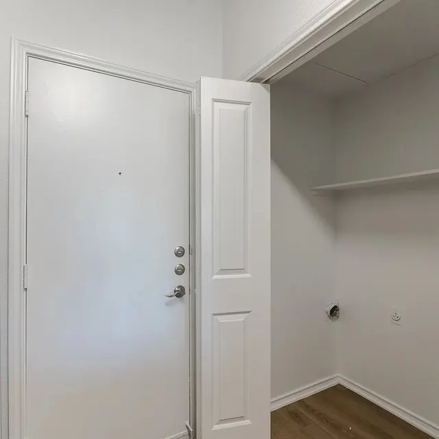 An entryway with an open door and a visible storage area on the right, featuring a shelf and space for appliances.