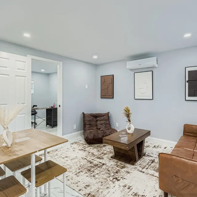 A modern living room featuring a brown sofa, a large coffee table, and a dining table with chairs. The walls are painted light blue, and there are abstract artwork pieces hanging on the walls. A door leads to another room, and a rug lies on the floor.