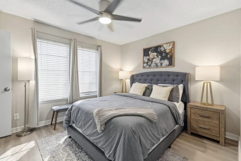 A cozy bedroom featuring a gray upholstered bed with decorative pillows, a bedside table with lamps, large windows covered with sheer curtains, and a modern floral wall art.