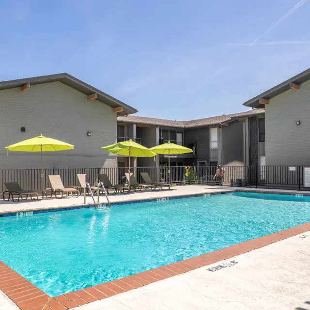 A bright swimming pool surrounded by recreational chairs and umbrellas at an apartment complex.