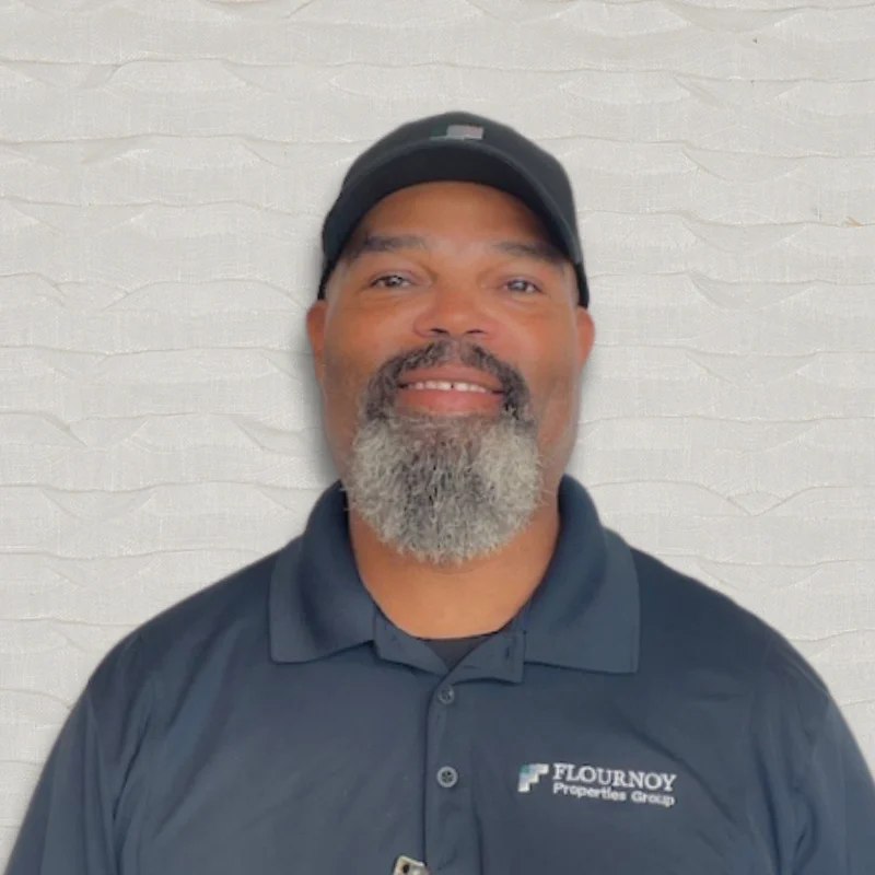 Portrait of a man with a beard and cap wearing a dark polo shirt with a logo, smiling against a textured light background.