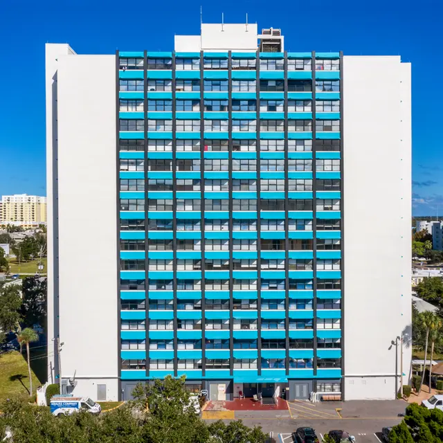Modern Residential Apartment Building A tall residential building featuring a modern design with blue and white exterior. The building has several balconies and is surrounded by greenery and parking spaces.