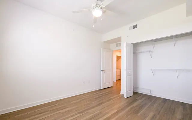 An empty room with light hardwood flooring, a ceiling fan, and a closet. A doorway leads to another room in the background.