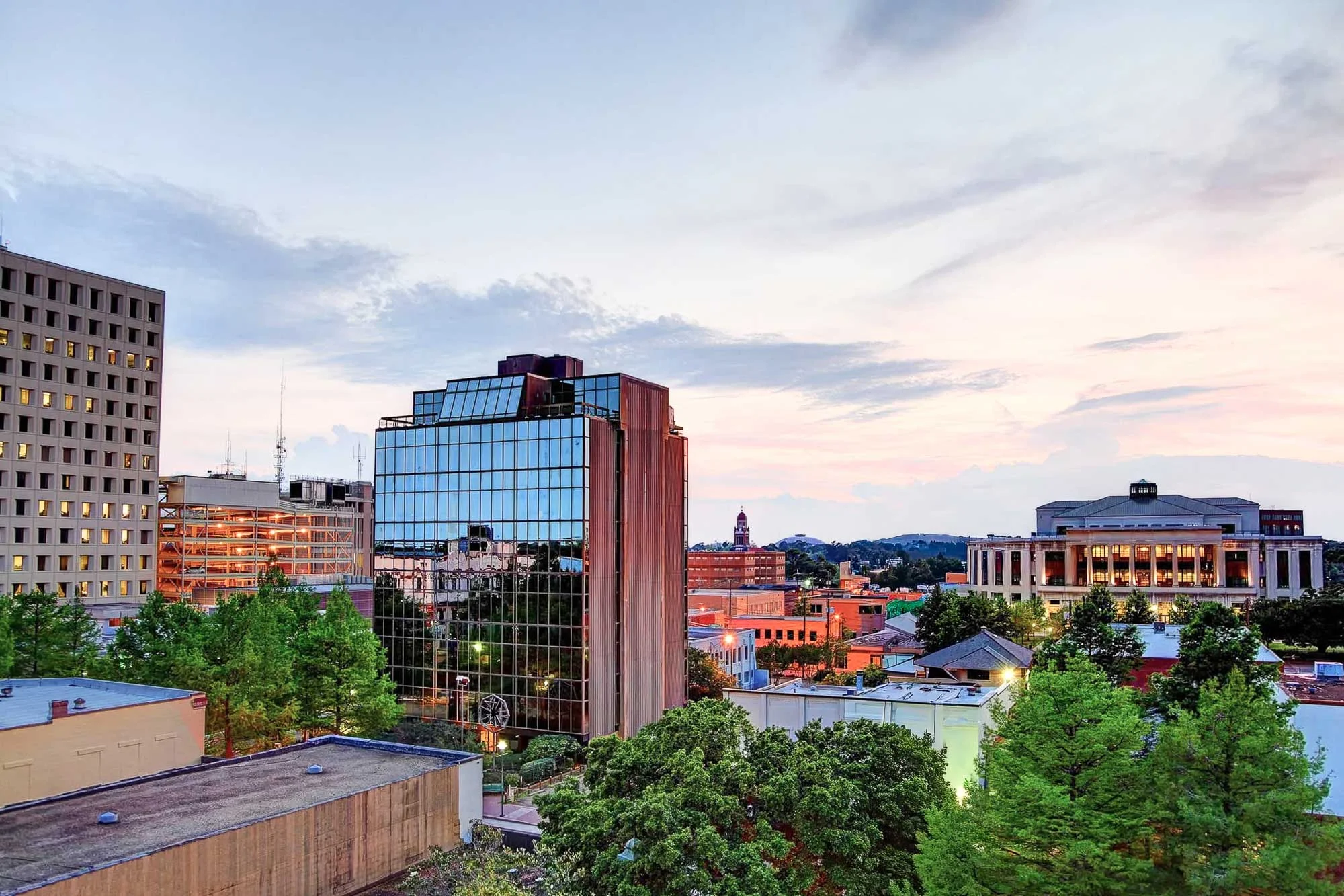 A skyline view of a city during sunset, featuring modern buildings and trees in the foreground with colorful clouds in the sky.