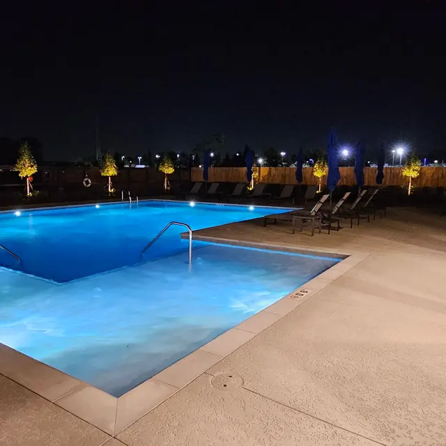 A well-lit swimming pool at night, surrounded by lounge chairs and illuminated trees, creating a serene atmosphere.