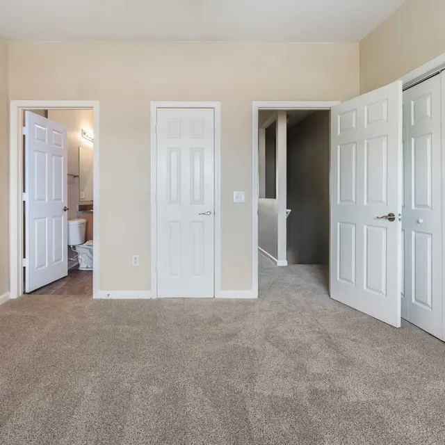 A spacious room featuring beige walls and light brown carpet. There are three white doors: one leads to a bathroom, while the others open to closets. The room is well-lit with natural light coming from the left side.